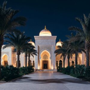 A grand white marble palace with illuminated domes and arches at night, viewed from a paved driveway lined with palm trees.