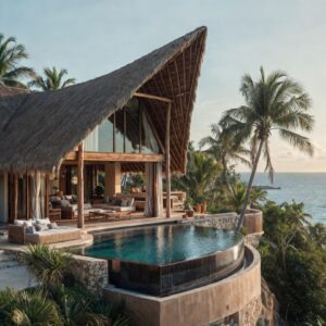 A grand tropical villa with high-pitched thatched roofs and a curved infinity pool overlooking a beach and a luxury yacht in the ocean.